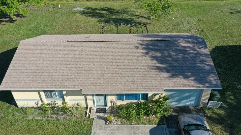 Aerial view of a large house with textured brown shingles, surrounded by manicured lawns and a few palm trees under a clear sky.