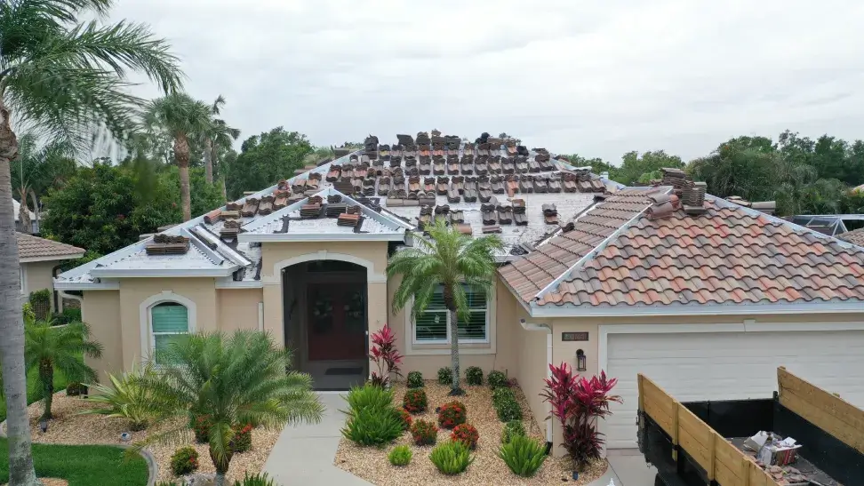 A large house with a severely damaged tile roof, surrounded by well-manicured gardens and neighboring homes.