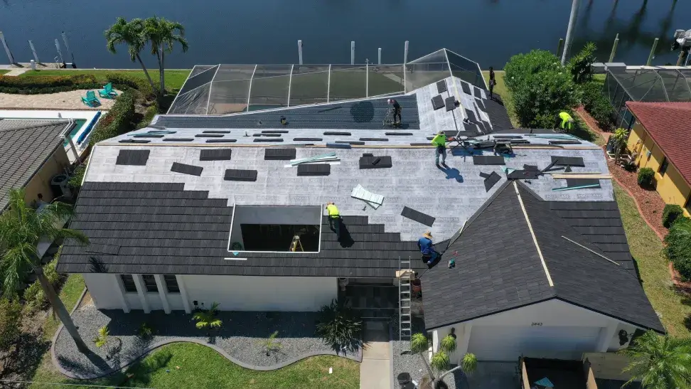 Aerial view of workers installing roofing shingles on a house near a water body.